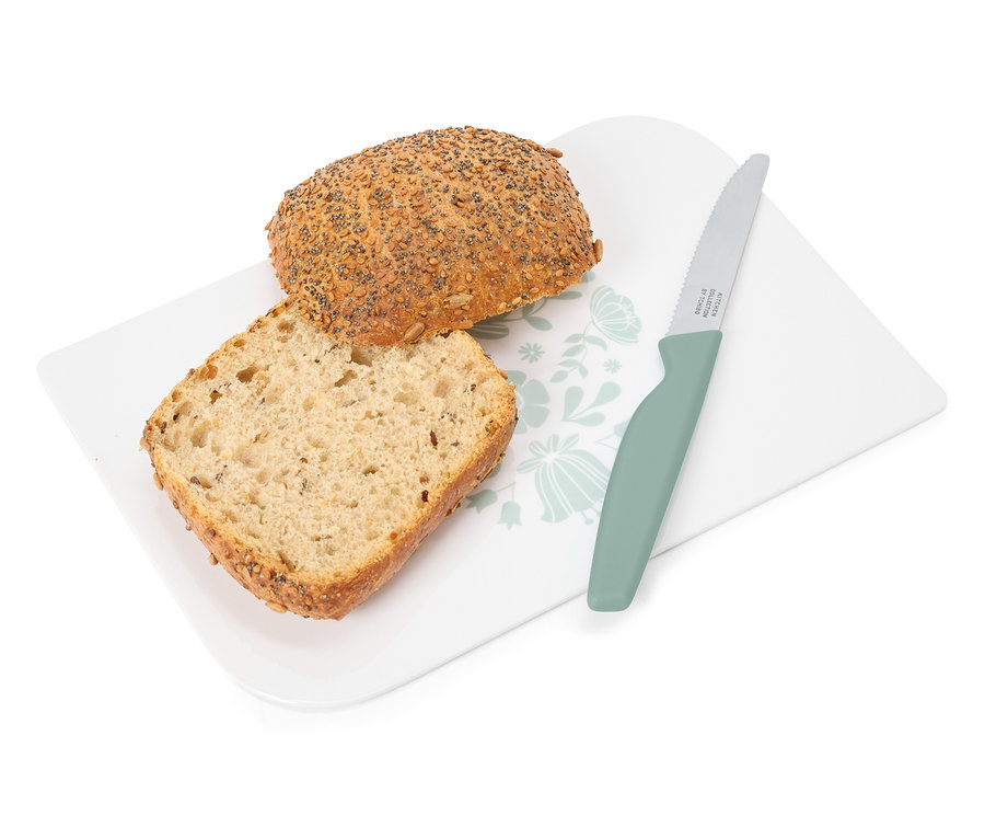 Auf einem weißen Brett mit Blumenmuster liegen eine Brotscheibe und ein ganzes Brötchen neben einem grünen Messer.