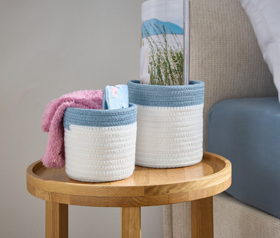 Zwei weiße geflochtene Körbe mit blauem Rand stehen auf einem Holztisch neben einem Bett.