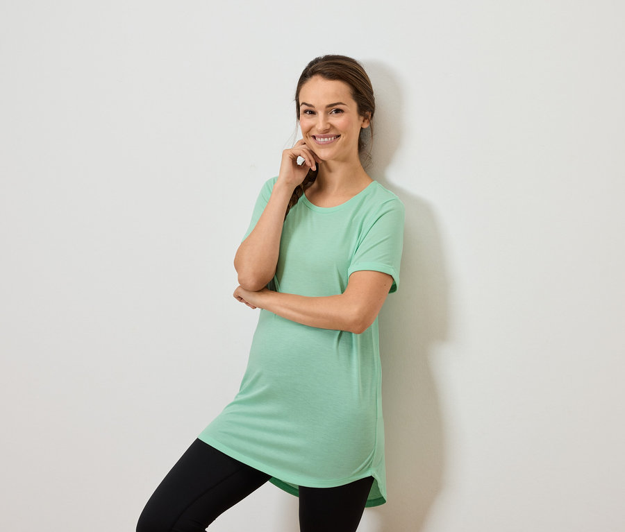 Frau in mintgrünem Oberteil und schwarzen Leggings posiert mit der Hand am Kinn.