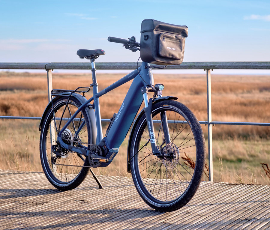 Blaues E-Bike mit FISCHER Lenkertasche PLUS+ Terra steht auf einem Holzsteg vor einer Dünenlandschaft.