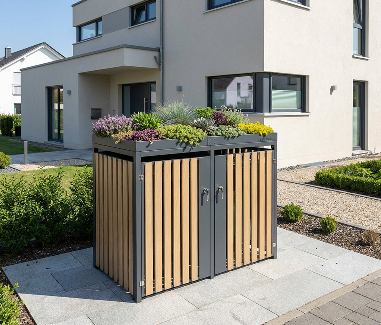 Eine graue Mülltonnenbox mit Blumenkasten, 2er, steht auf einer Terrasse vor einem Haus. Auf der Box befinden sich verschiedene Pflanzen.