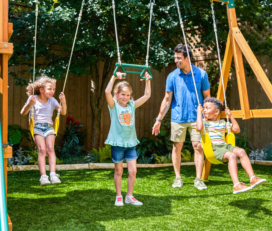 Vater steht neben drei Kindern, die auf einem Spielturm »Bolzo« im Garten spielen.