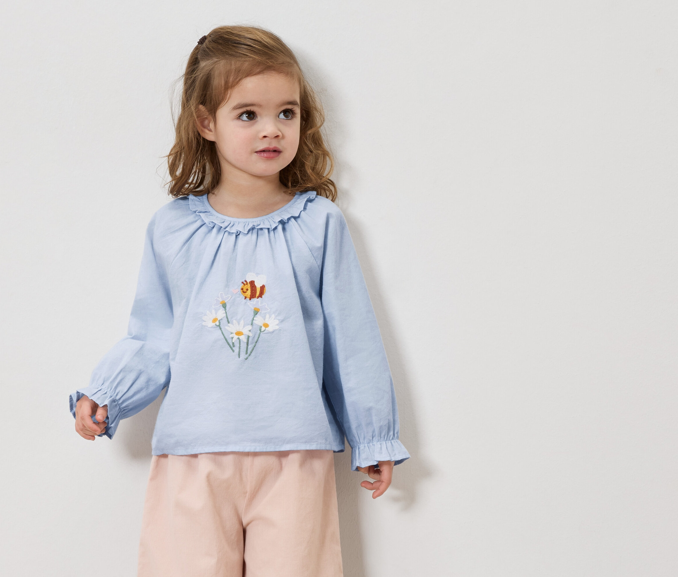 Mädchen mit blonden Haaren trägt eine hellblaue Bluse mit Gänseblümchen- und Bienenstickerei und eine rosafarbene Hose.