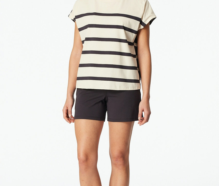 Frau steht barfuß in einem gestreiften Oberteil und schwarzen Shorts vor einem weißen Hintergrund.