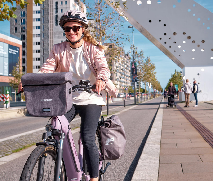 Eine Frau fährt mit einem lila Fahrrad mit der FISCHER Lenkertasche PLUS Cita durch die Stadt.