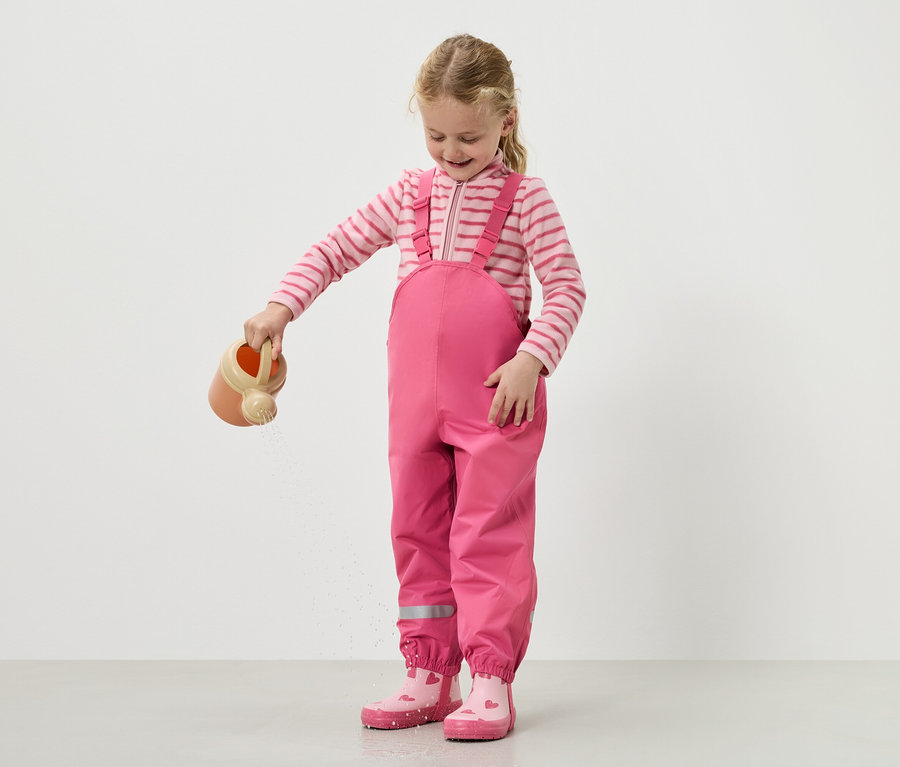 Ein Mädchen gießt Wasser mit einer Gießkanne, während sie eine rosa Latzhose und Stiefel trägt.