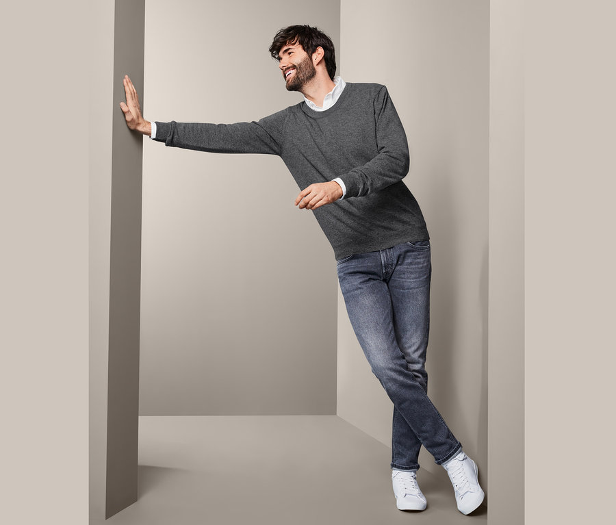 Mann lehnt an Wand und trägt Cashmere-Pullover mit Rundhalsausschnitt, Jeans und Turnschuhe.