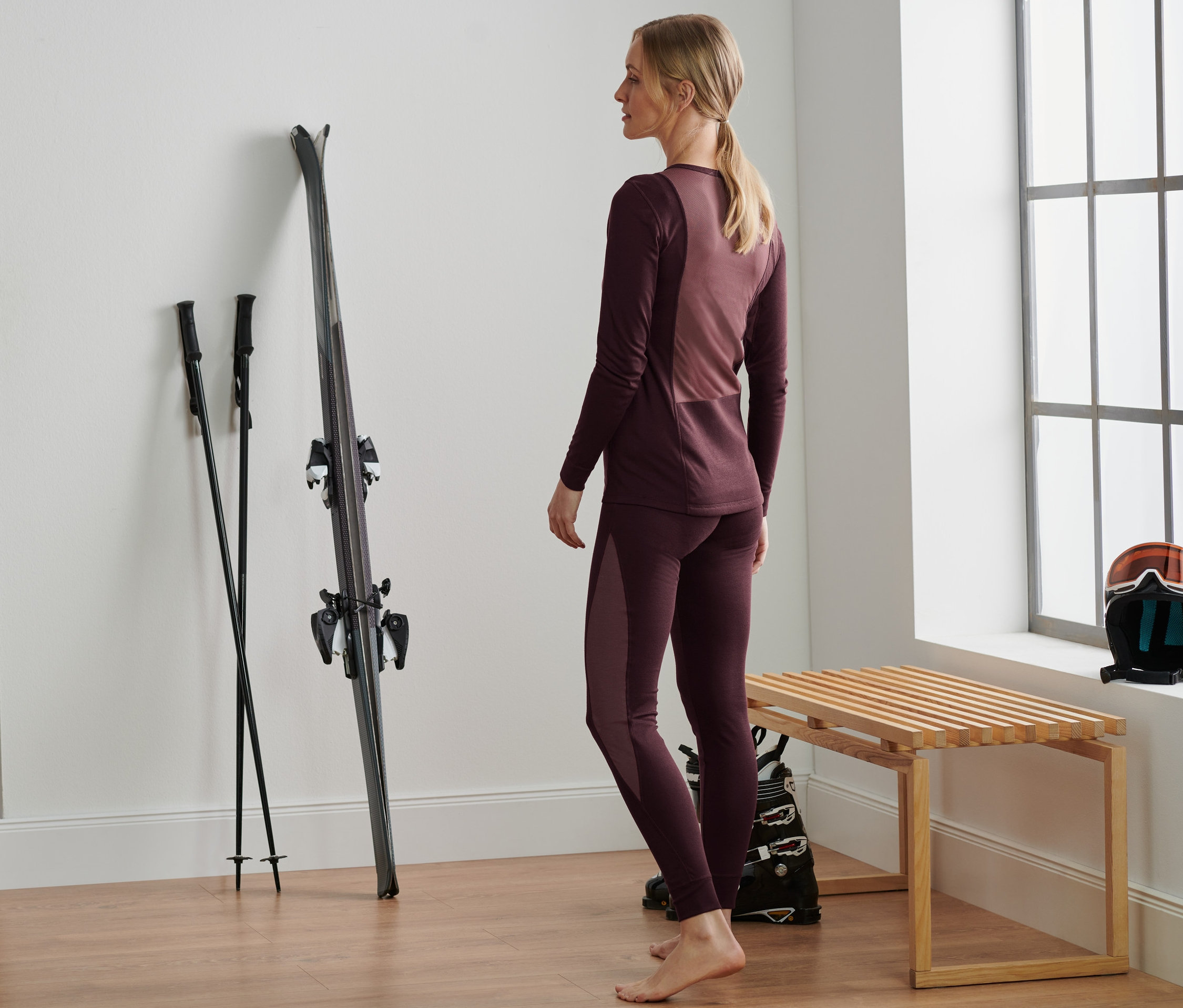 Frau steht in bordeauxfarbener Thermo-Funktionsunterwäsche neben Skiern und Stöcken.