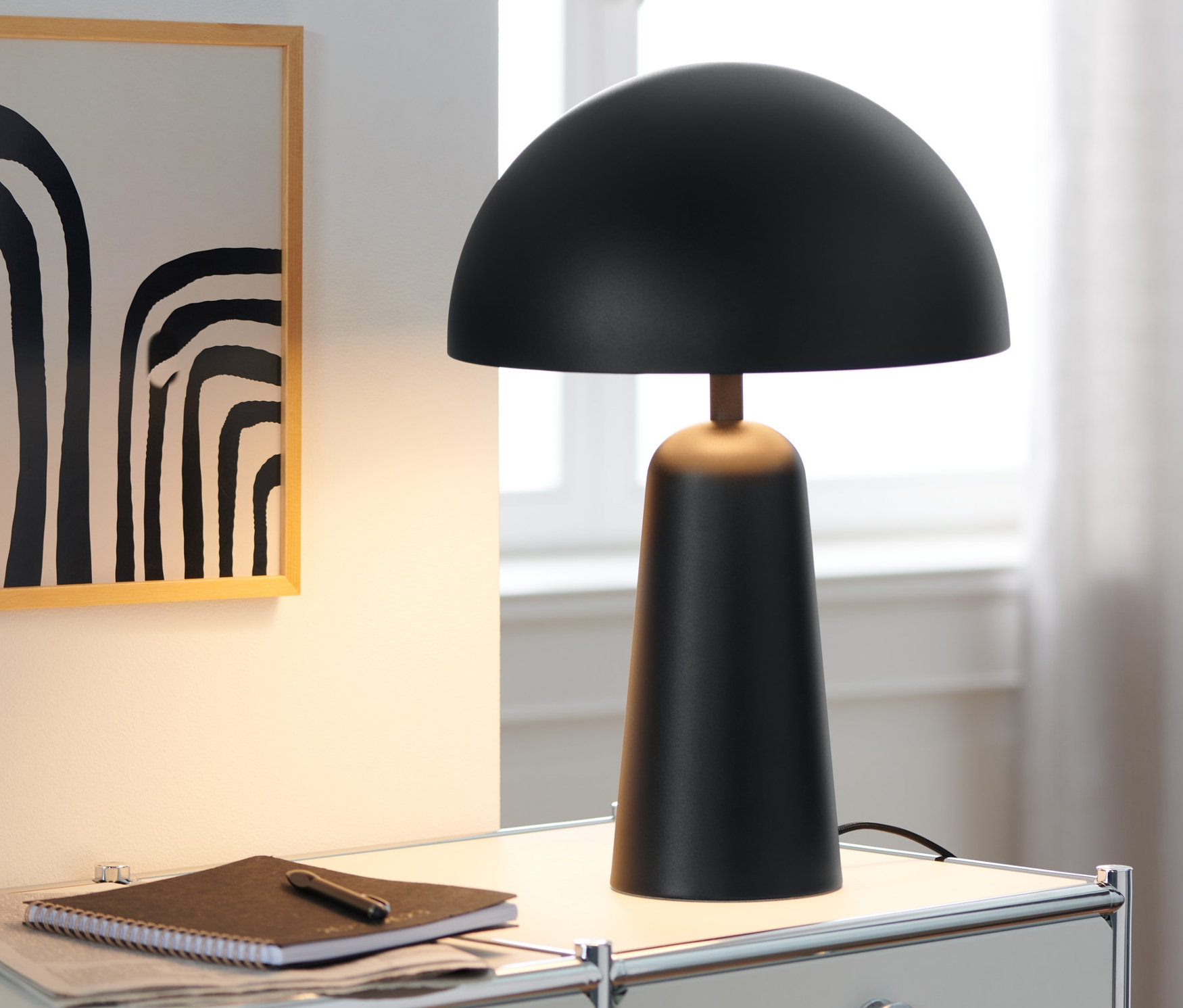 Eine schwarze Tischleuchte mit Pilzkopf steht auf einem weißen Tisch neben einem Notizbuch und einem Stift.