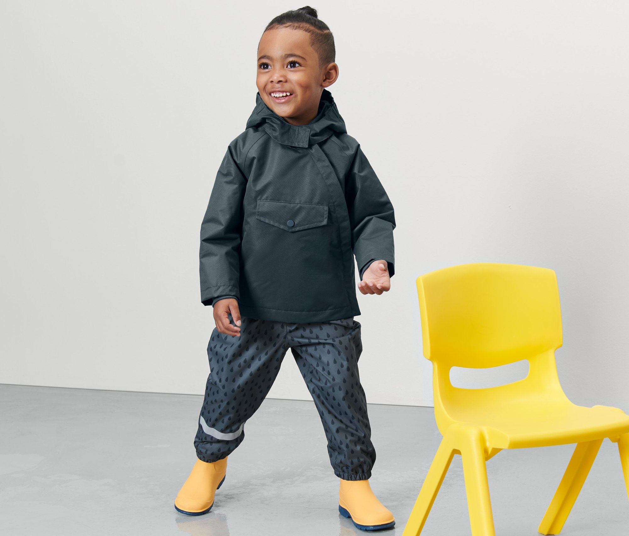 Ein Junge steht neben einem gelben Stuhl und trägt eine schieferblaue Kleinkind Thermo-Regenjacke.