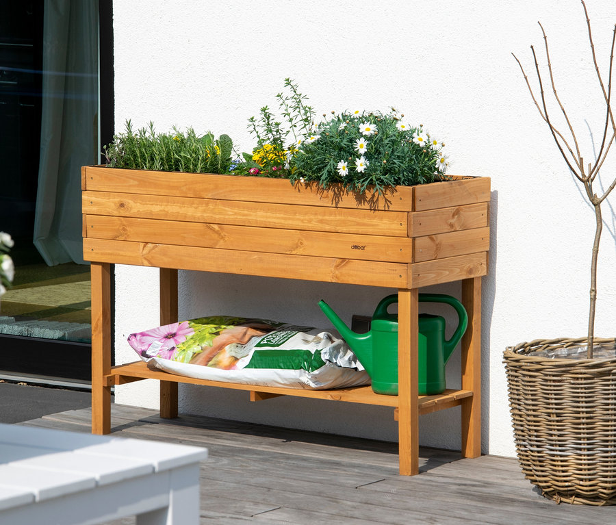 Ein hölzernes Hochbeet mit Pflanzen, daneben ein Blumentopf und eine Gießkanne.