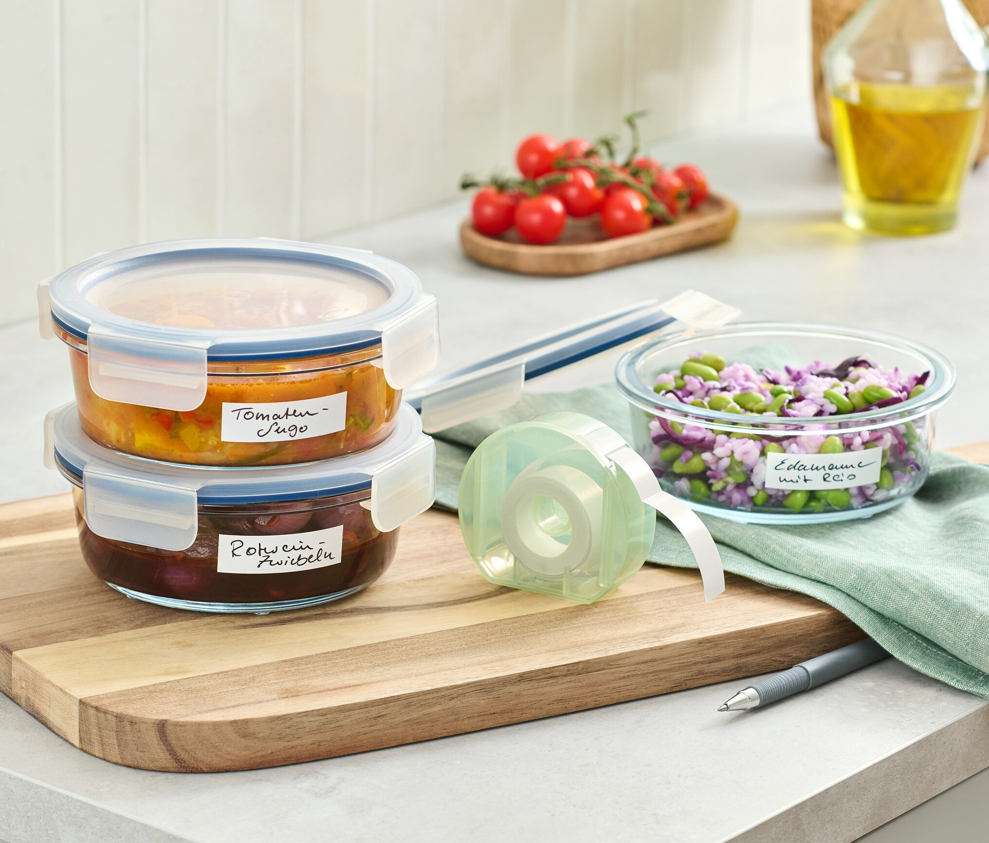 Zwei Glasbehälter mit Deckel gestapelt neben einem Etikettenspender und einer Schüssel mit Lebensmitteln.