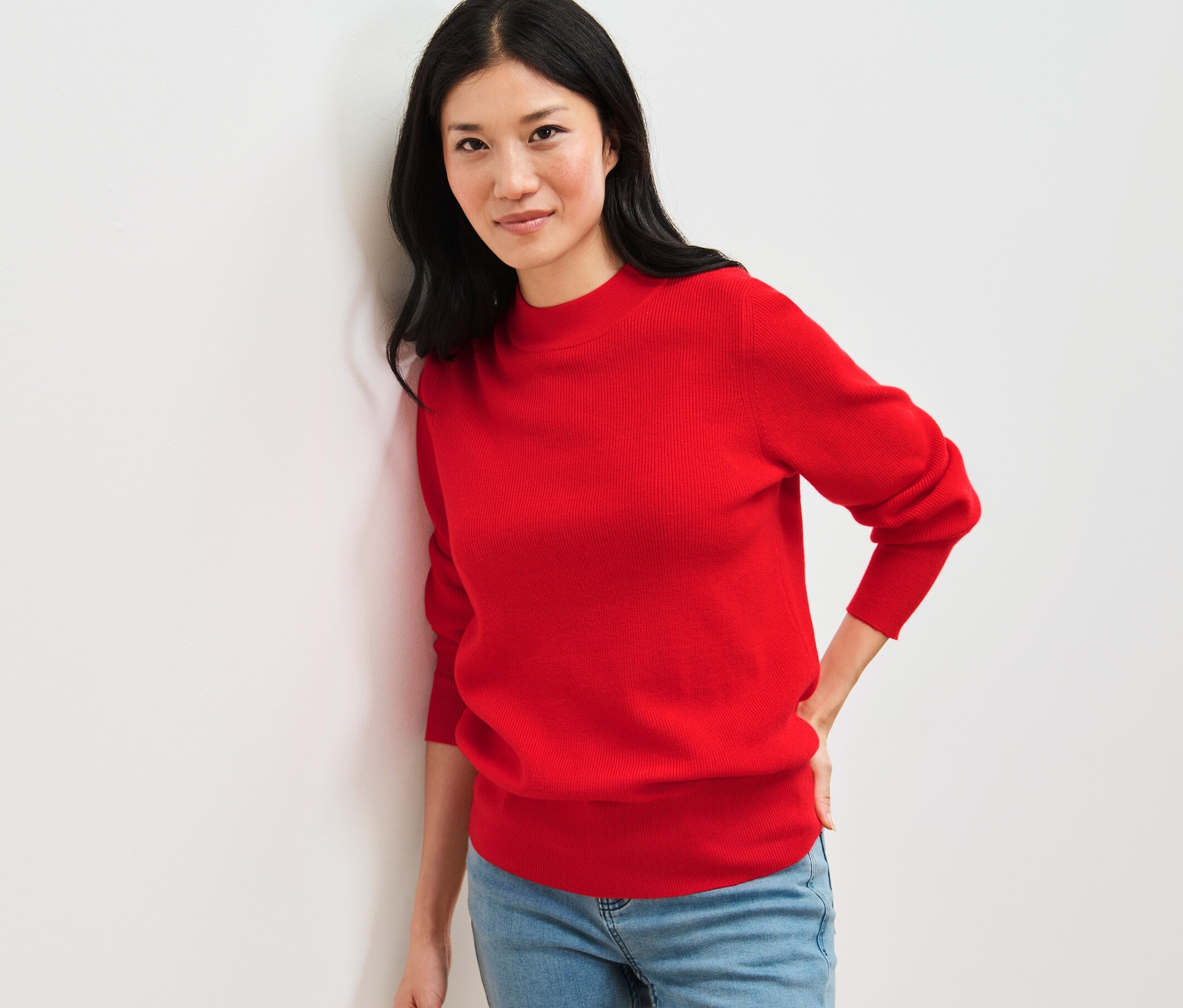 Frau lehnt an Wand und trägt roten Feinstrickpullover und Jeans.