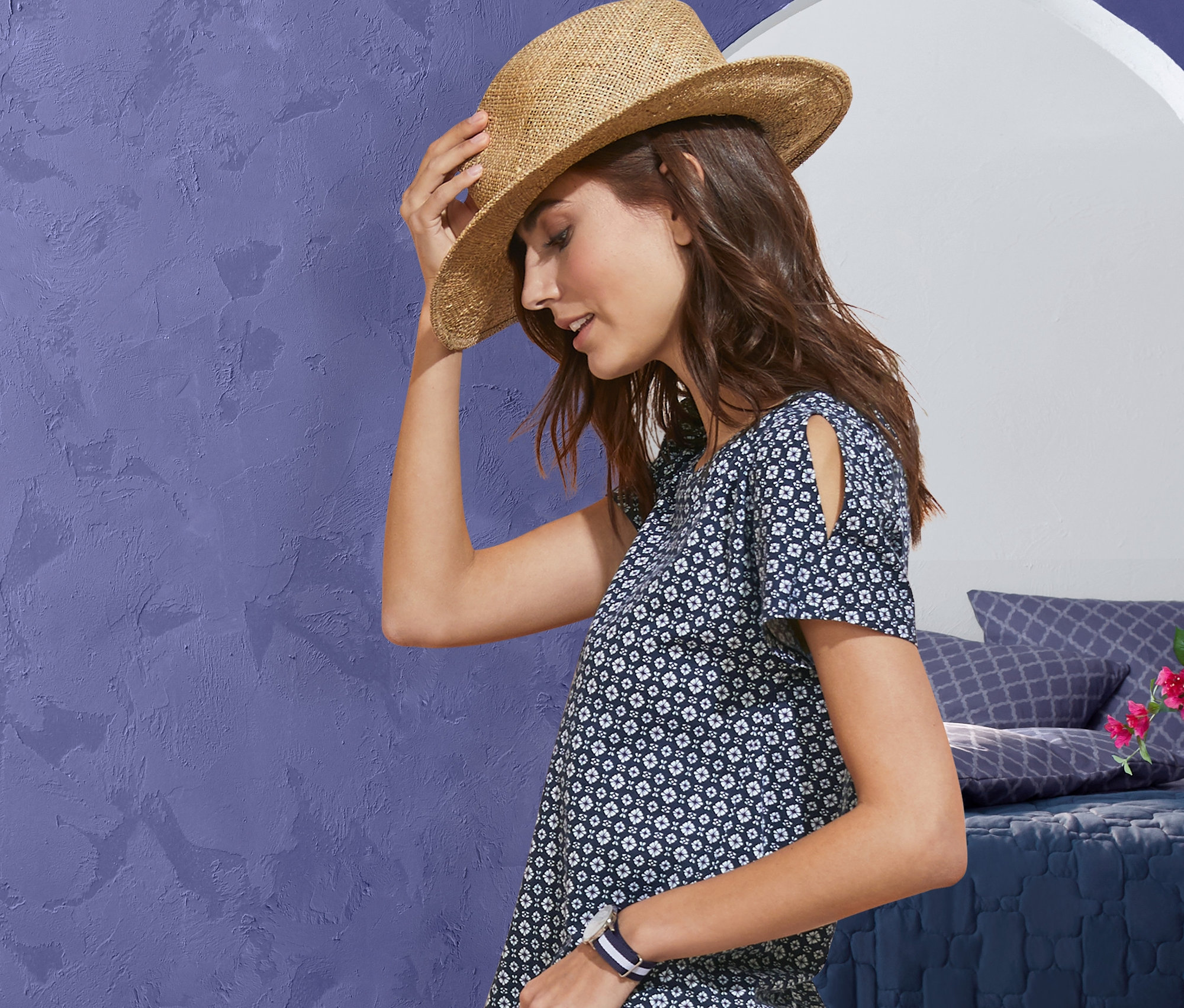 Frau hält einen Strohhut, trägt ein Shirt mit Alloverprint und eine Stretch-Jeans in 7/8-Länge. Im Hintergrund sind eine Bettwäsche, eine Tagesdecke und ein Sitzpouf zu sehen.