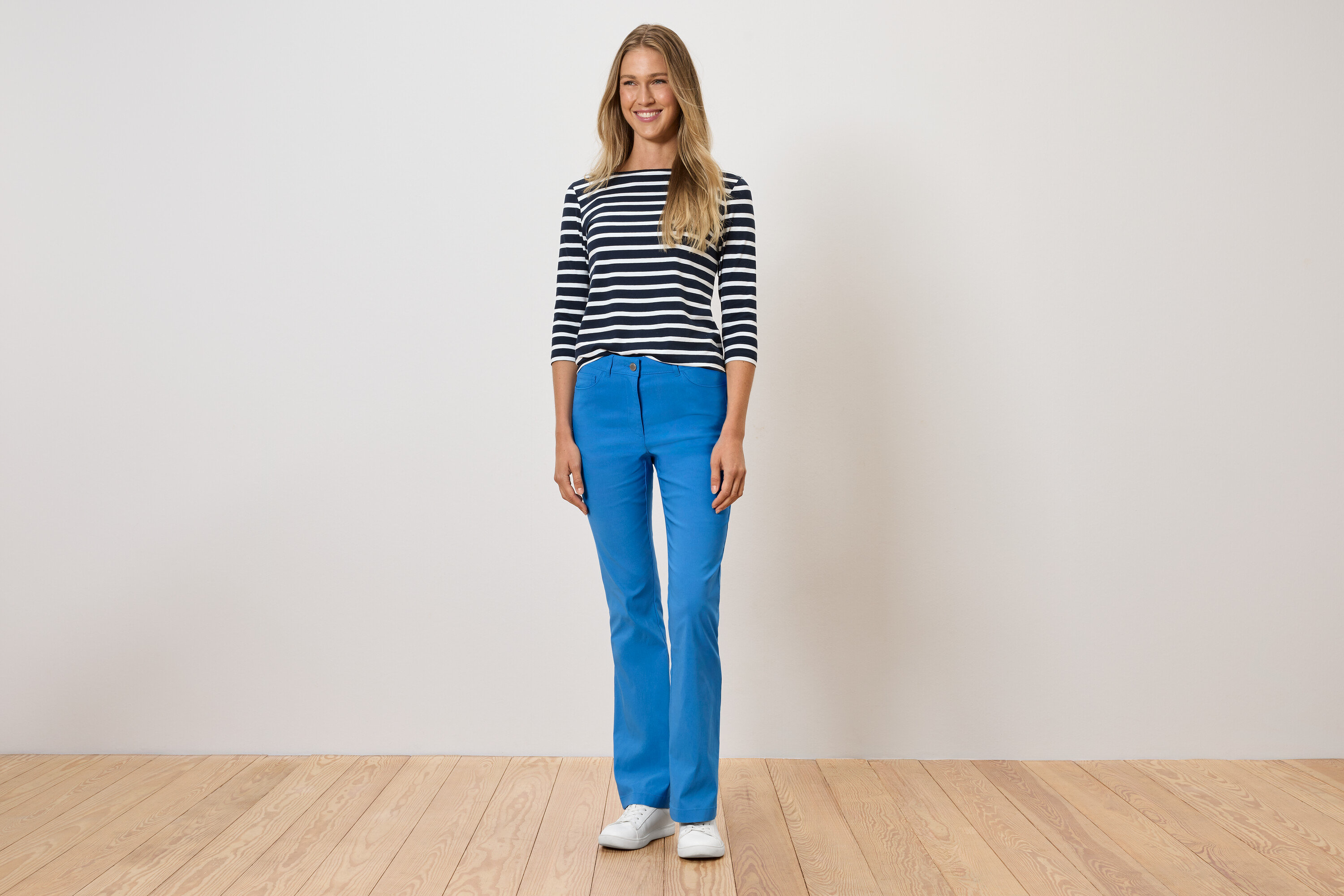 Frau steht und trägt blaue Hosen, ein gestreiftes T-Shirt und weiße Schuhe.
