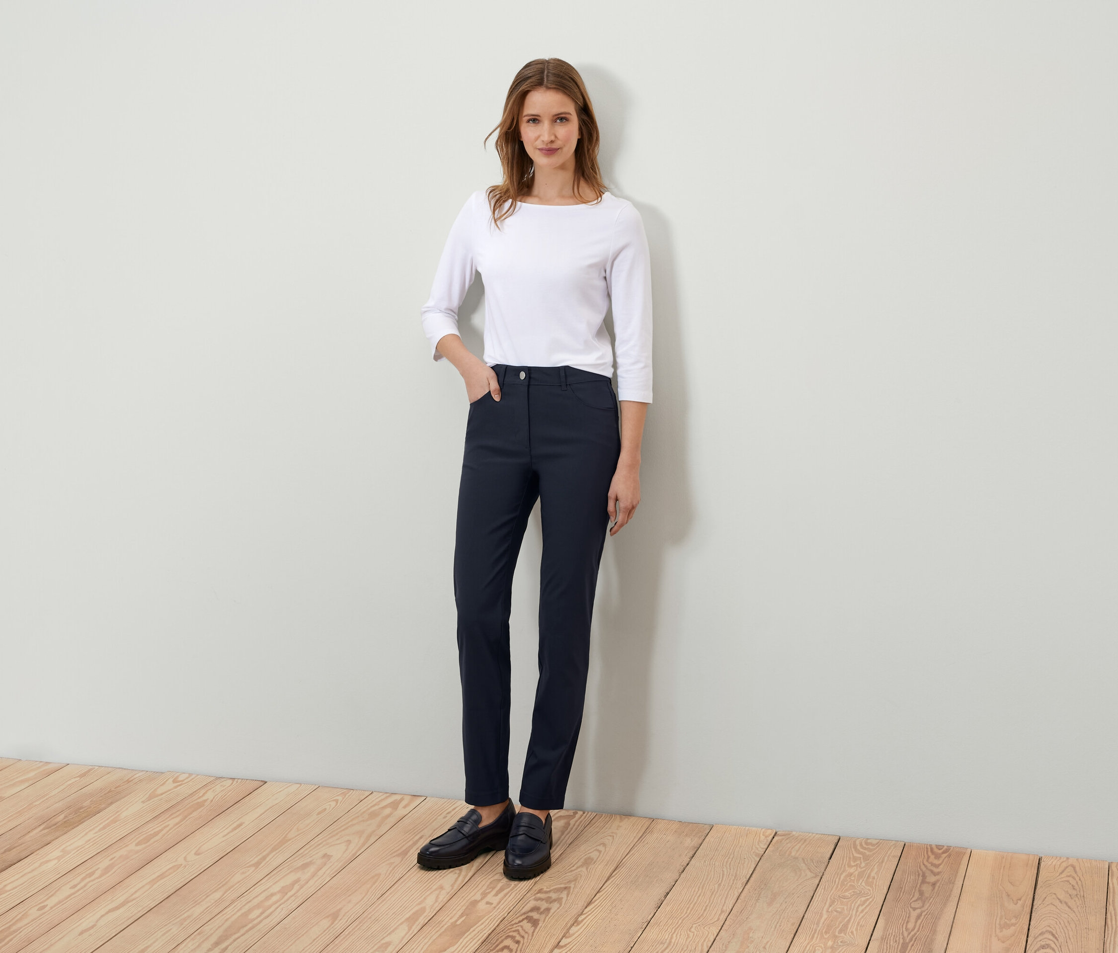 Frau steht und posiert in einem weißen Dreiviertelarm-Shirt und dunkelblauen Hosen.