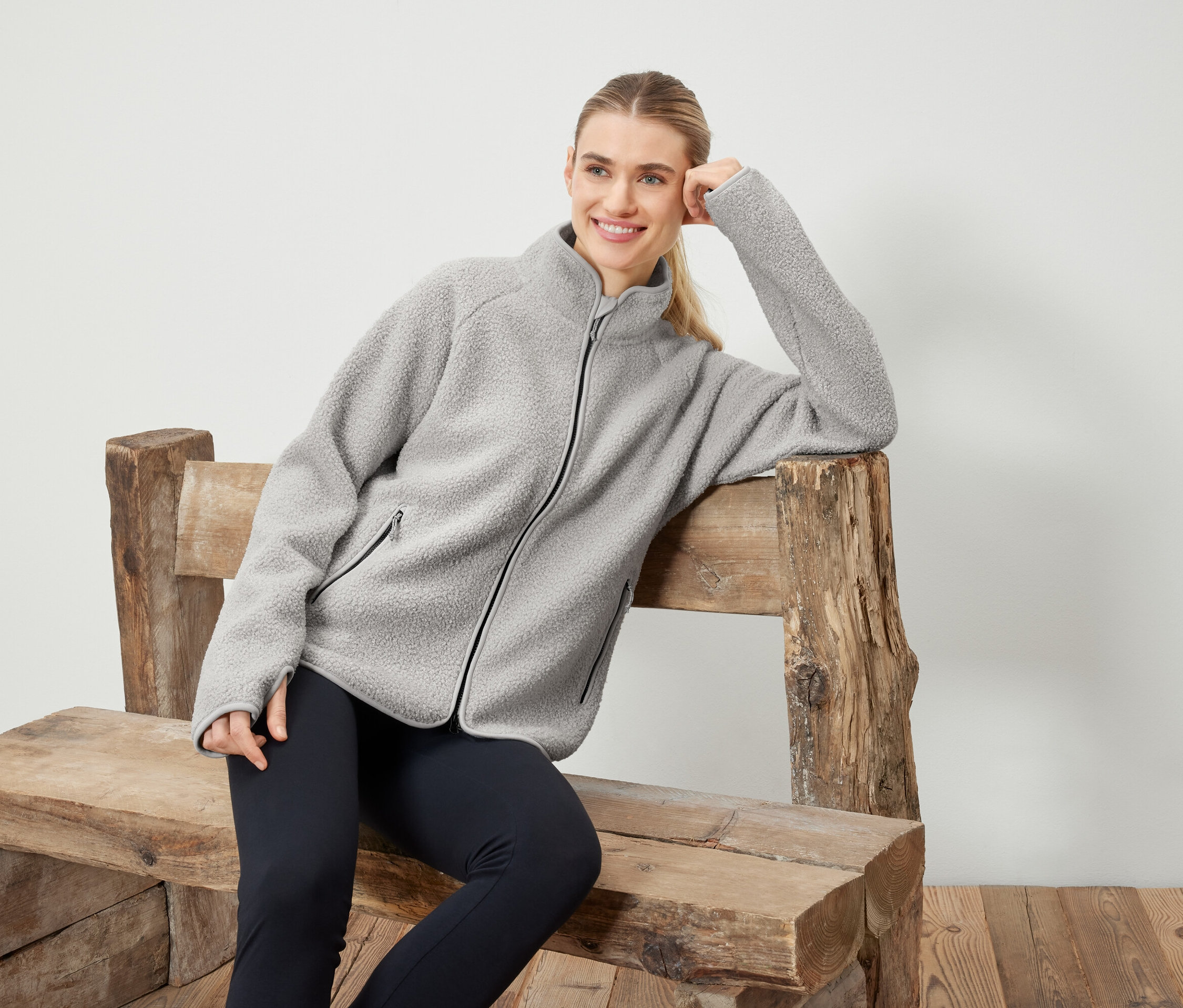 Frau in grauer Fleecejacke posiert auf einer Holzbank.