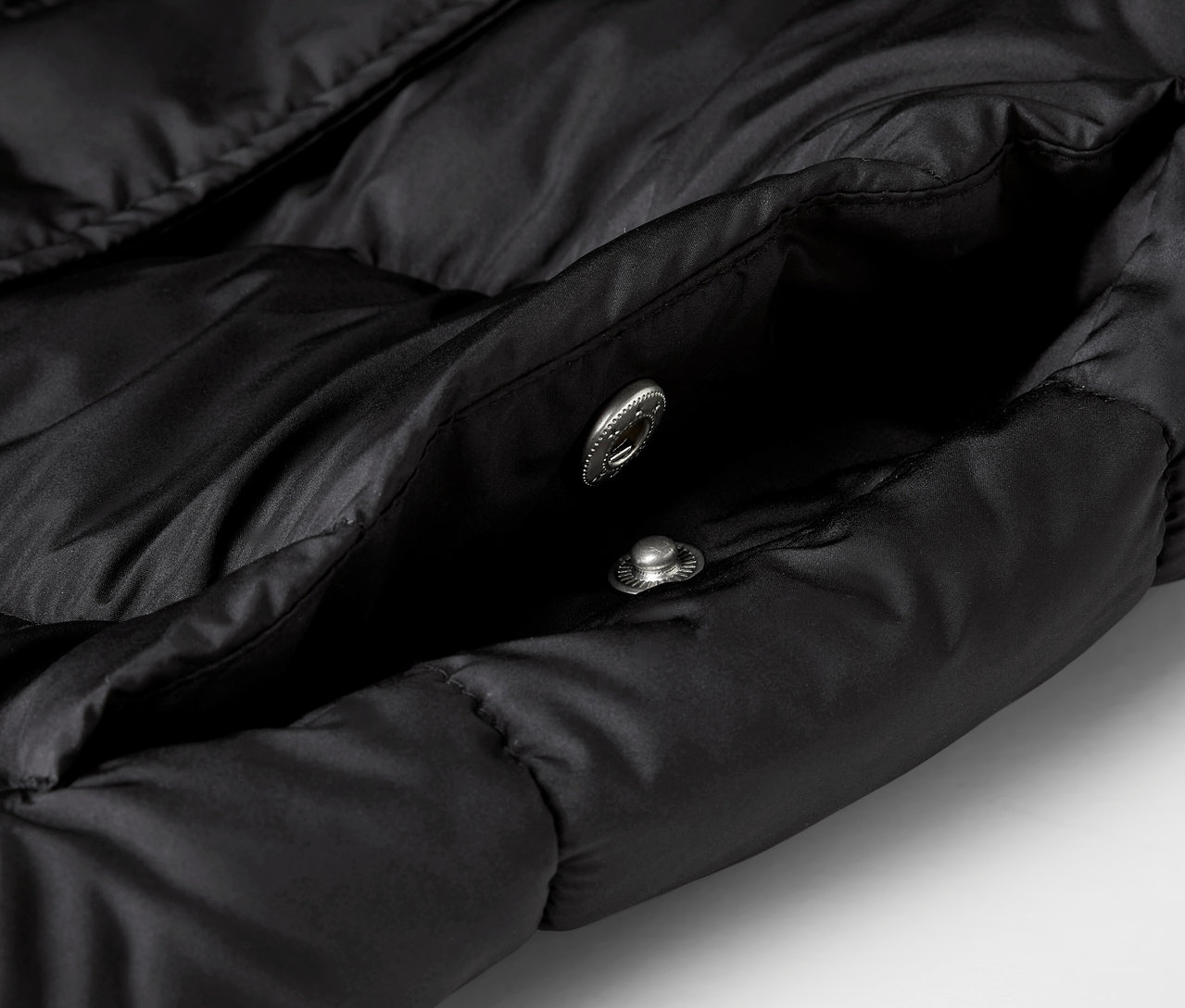 Detailaufnahme einer schwarzen Steppjacke mit offener Tasche und zwei silbernen Druckknöpfen.