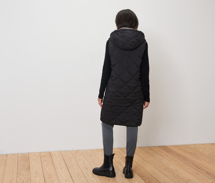 Frau mit dem Rücken zur Kamera trägt eine gesteppte schwarze Weste mit Kapuze, schwarze Stiefel und graue Leggings.