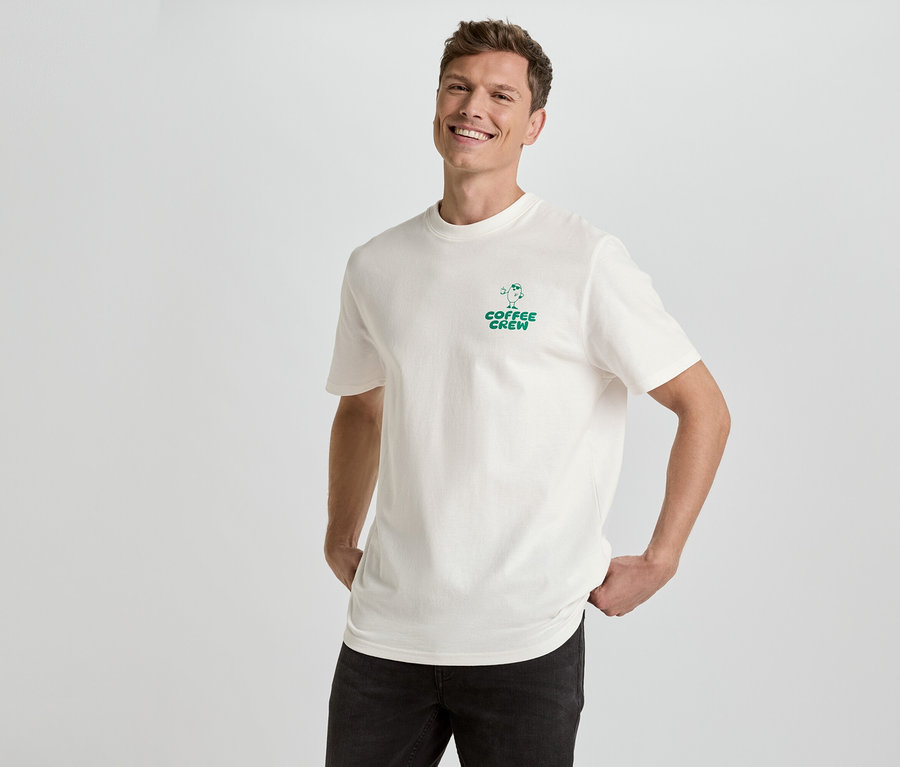 Mann lächelt und posiert in einem cremefarbenen T-Shirt mit dem grünen Aufdruck "Coffee Crew" und schwarzen Jeans vor einem weißen Hintergrund.