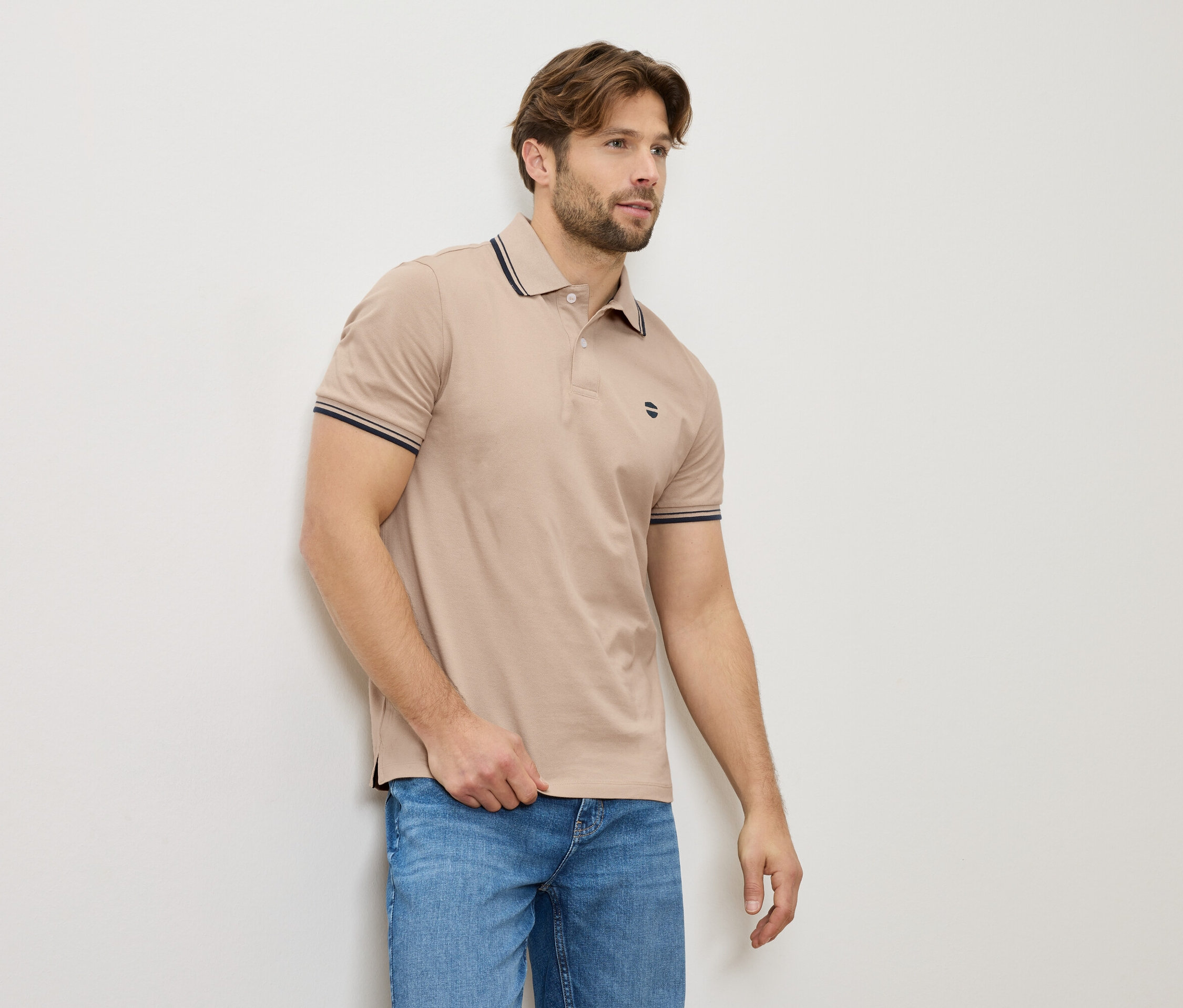 Ein Mann in einem beigen Poloshirt und Jeans steht an einer Wand.