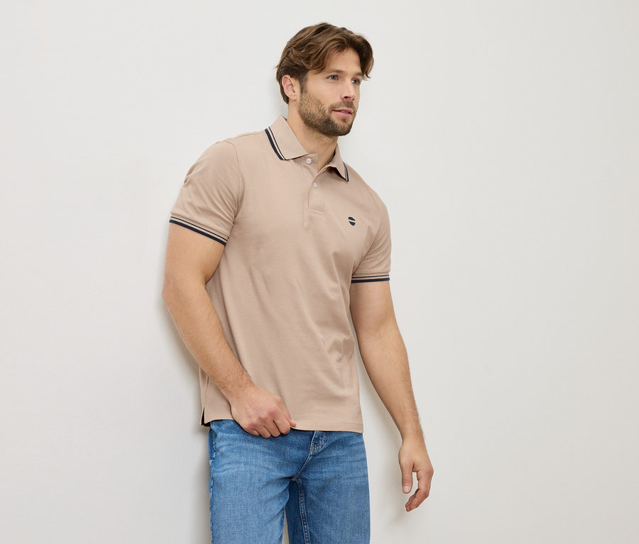 Ein Mann in einem beigen Poloshirt und Jeans steht an einer Wand.