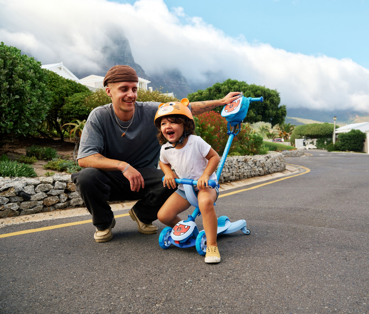 Ein Mann kniet neben einem Kind auf einem blauen Hudora 4-in-1-Scooter, Cyan. Das Kind trägt einen orangefarbenen Helm mit Ohren.
