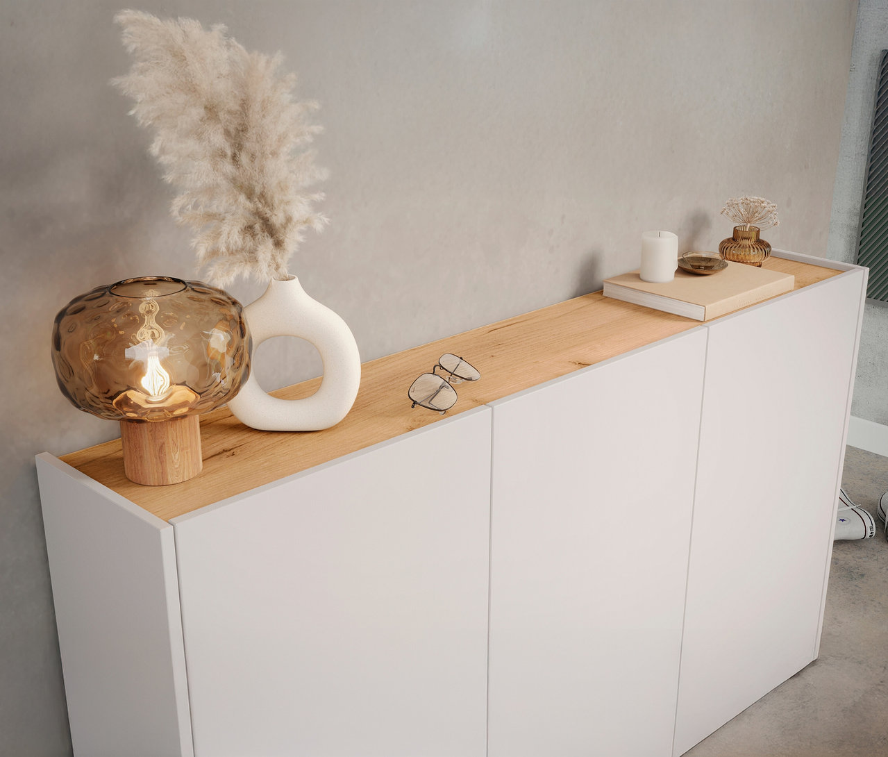 Auf einem weißen Schrank mit Holzplatte stehen eine braune Lampe, eine Vase mit Pampasgras, eine Brille, eine Kerze und ein Buch.