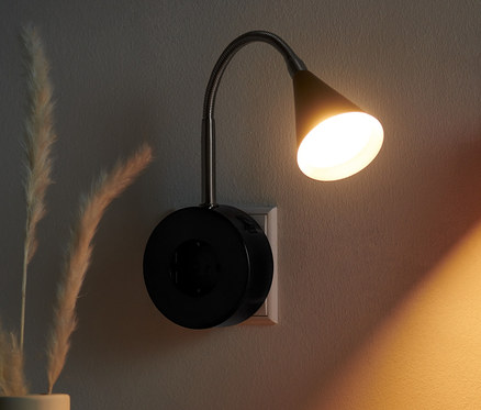 Aufnahme eines Nachttisches mit Buch, Kerze und Vase mit Gras, an der Wand eine LED-Schwenkleuchte.