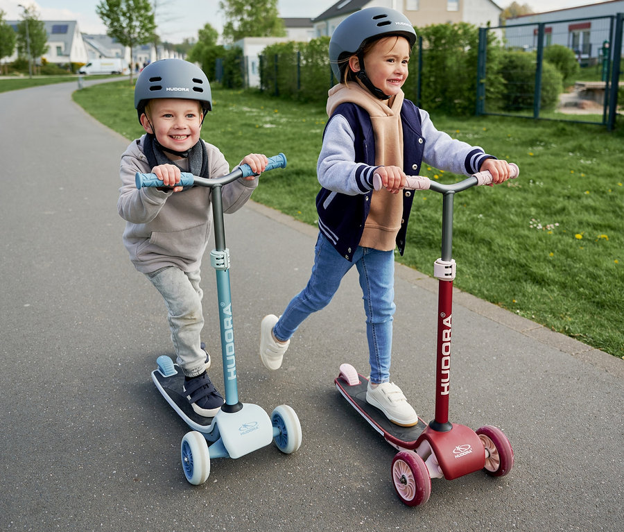 Kinder fahren mit HUDORA Tri-Scootern, einer bordeauxrot, der andere blau.