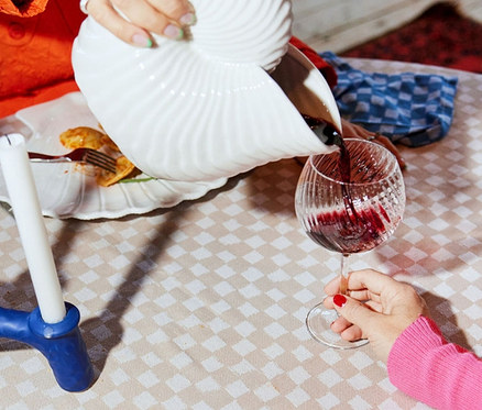 Person schenkt Wein aus einem muschelförmigen Krug in ein Weinglas auf einem Tisch mit Speisen.