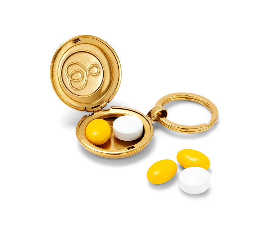 Geöffnete goldene Pillendosen-Schlüsselbund mit einer gelben und einer weißen Pille, zwei Pillen liegen daneben.