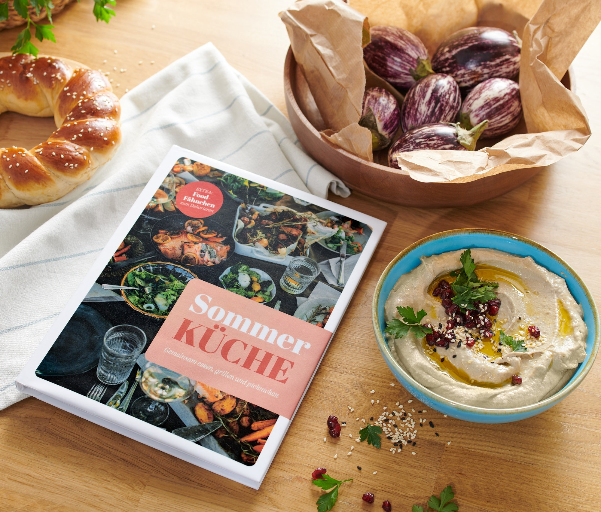 Auf einem Holztisch liegen das Buch »Sommerküche«, ein Kranzkuchen, eine Schale mit Auberginen und eine Schale mit Hummus.