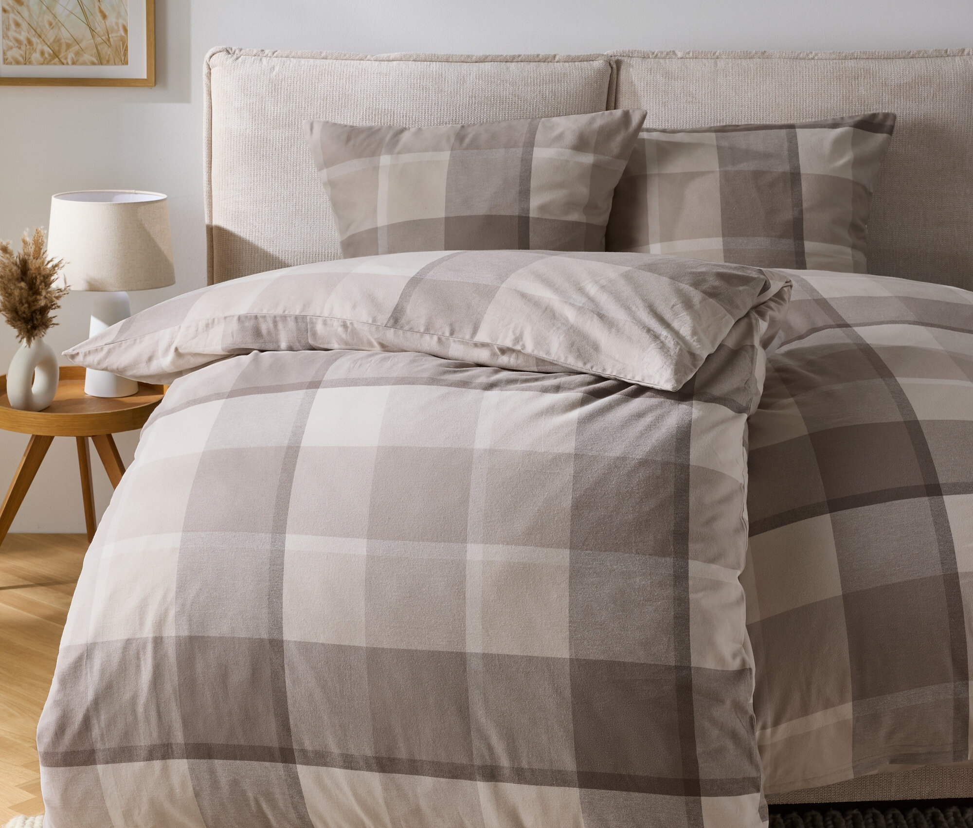 Ein kariertes Bettwäsche-Set auf einem Bett mit Kopfteil, auf einem Holzboden liegt ein cremefarbener Teppich.