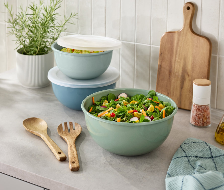 Auf einer Küchentheke stehen eine Salatschüssel, ein Holzbesteck, ein Salzstreuer, eine Ölflasche, ein Schneidebrett und eine Topfpflanze.