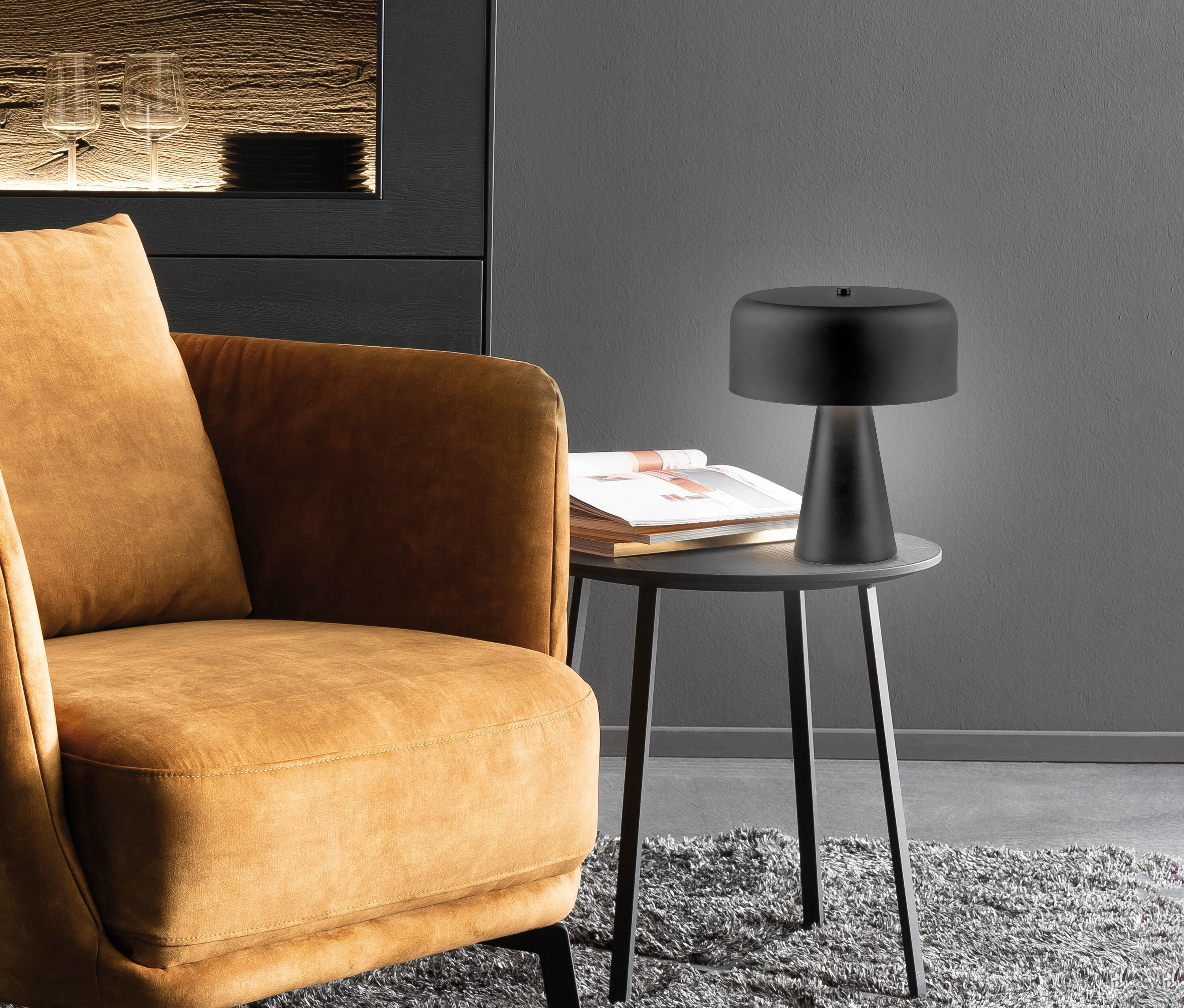 Ein brauner Sessel steht neben einem runden Tisch mit einer schwarzen Lampe und einem Buch.