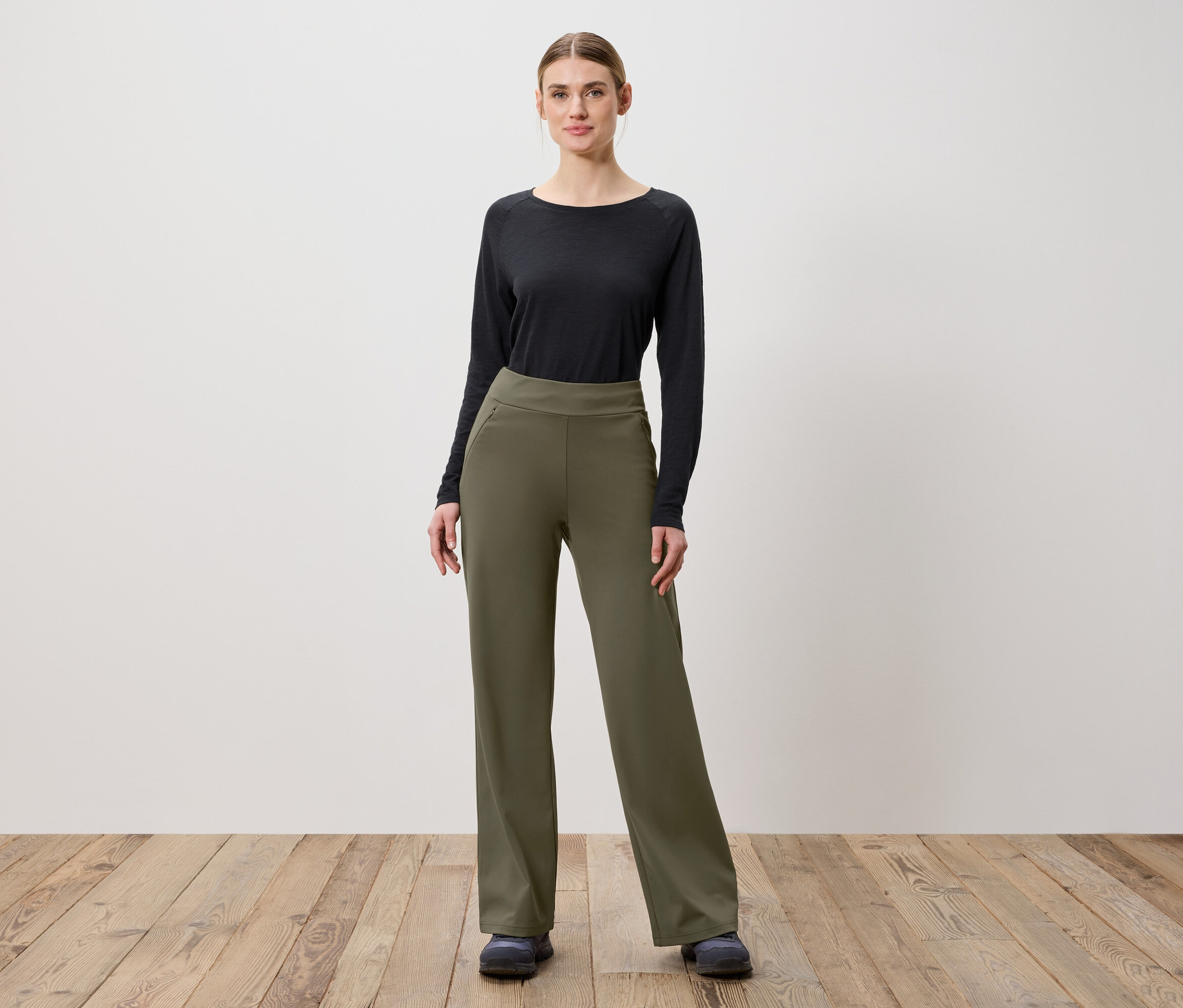 Eine Frau posiert auf einem Holzboden in olivgrünen Hosen und einem schwarzen Langarmshirt.