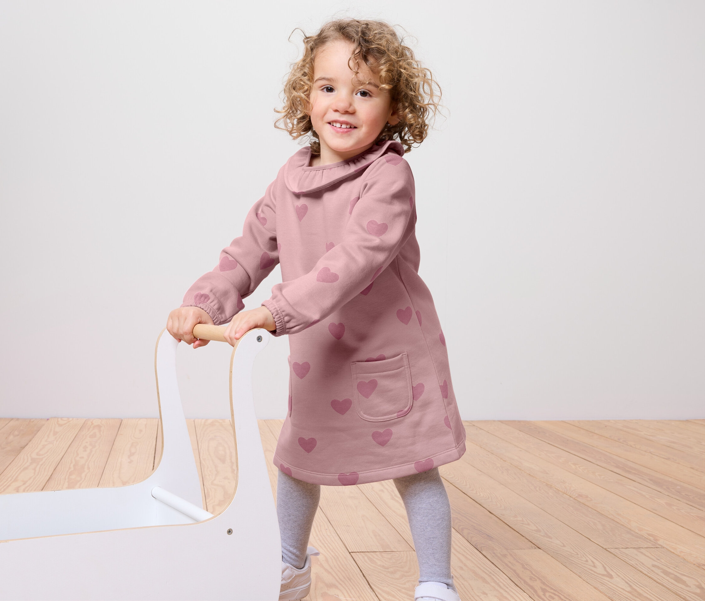 Ein lächelndes kleines Mädchen schiebt einen weißen Spielzeugwagen. Sie trägt ein rosa Kleid mit Herzen, graue Leggings und weiße Turnschuhe.