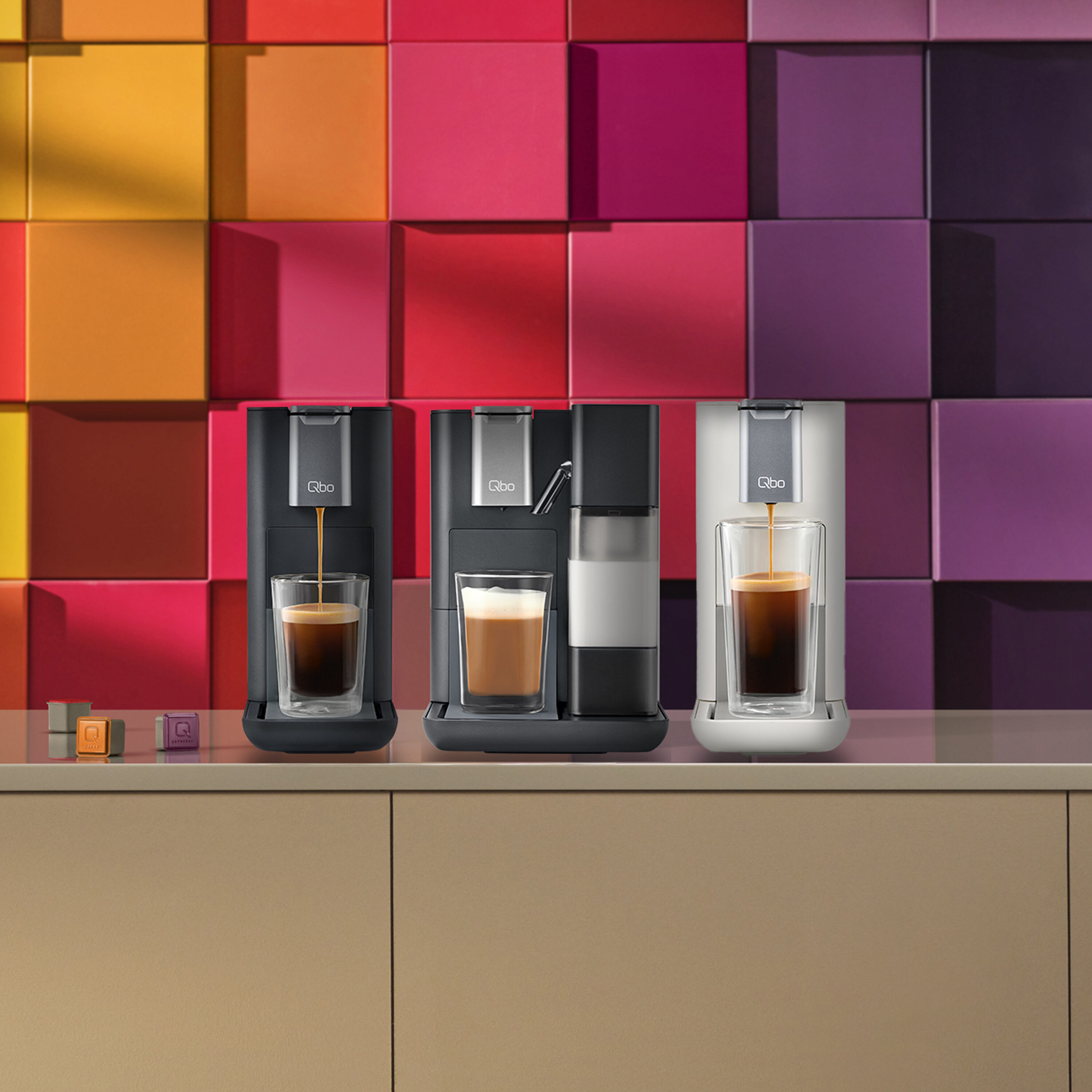 Drei Qbo Kaffeemaschinen in Schwarz und Weiß stehen auf einer Theke, während Kaffee in Gläser gefüllt wird.