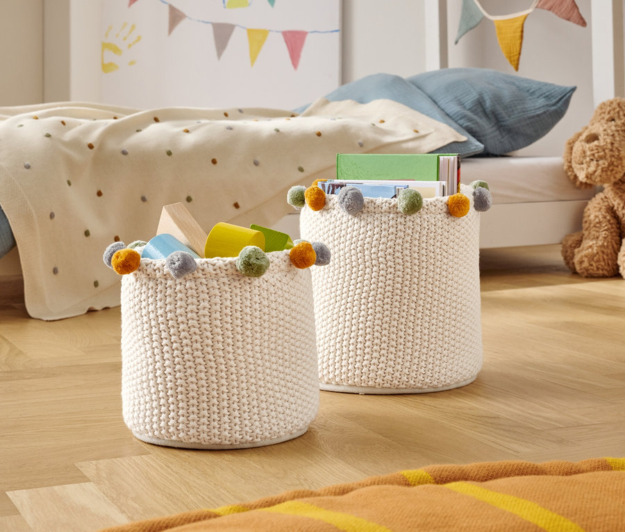 Zwei gestrickte Aufbewahrungskörbe mit bunten Bommeln stehen auf einem Holzboden in einem Kinderzimmer.