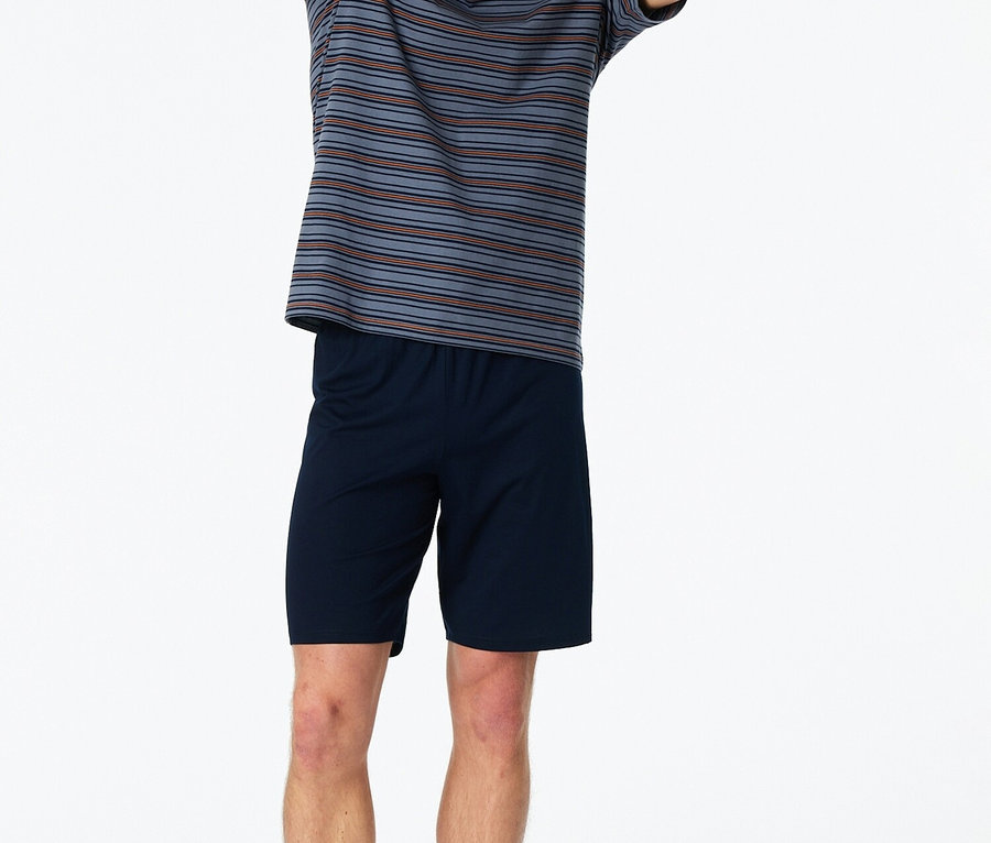 Mann steht barfuß mit erhobenen Händen im gestreiften T-Shirt und blauen Shorts vor einem weißen Hintergrund.