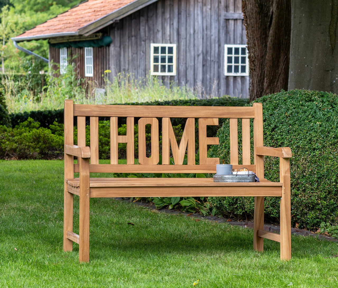 Eine deVries Gartenbank »Home« steht im Garten vor einem kleinen Haus.