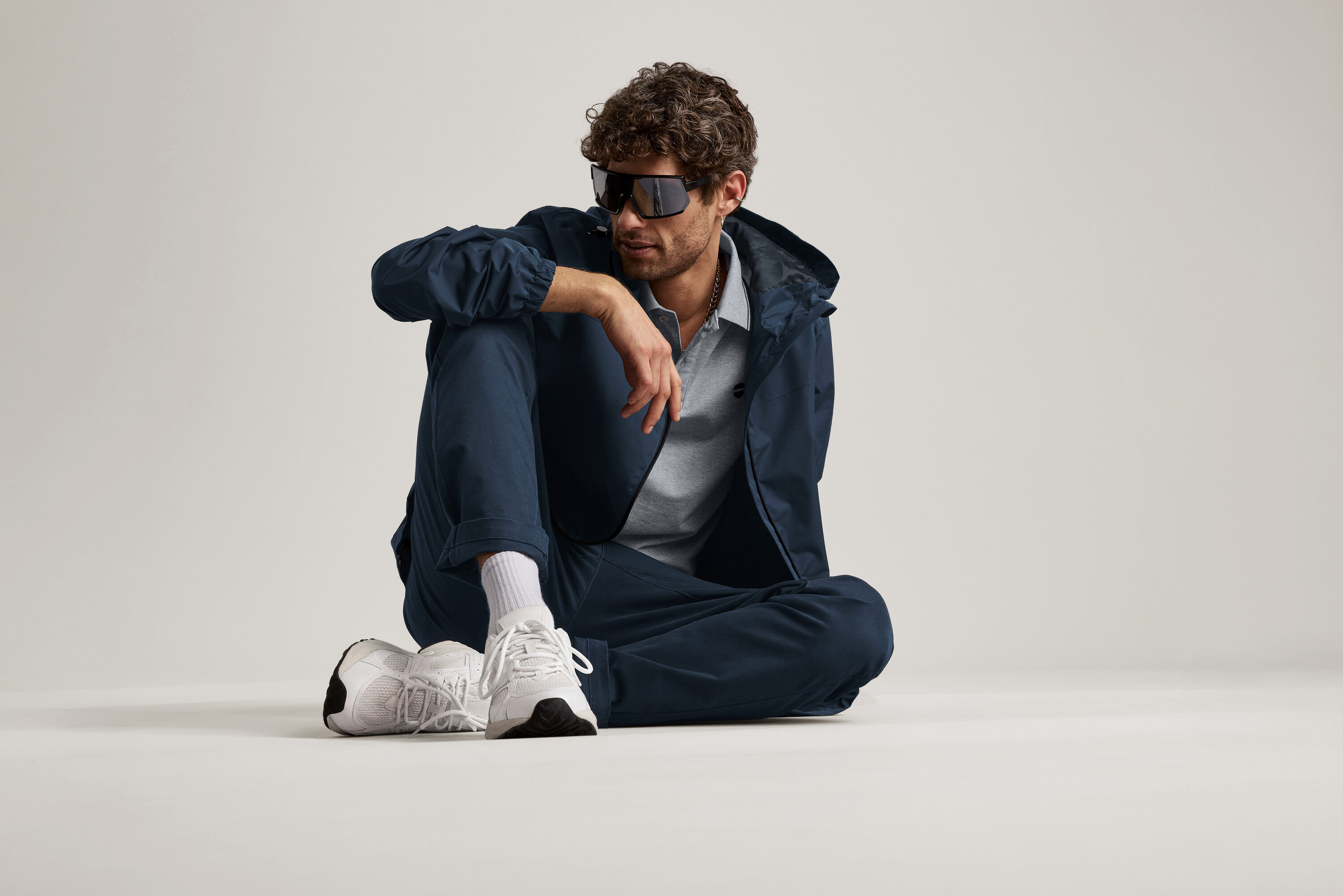 Ein Mann sitzt auf dem Boden und trägt eine marineblaue Chino, eine Jacke und eine Sonnenbrille.