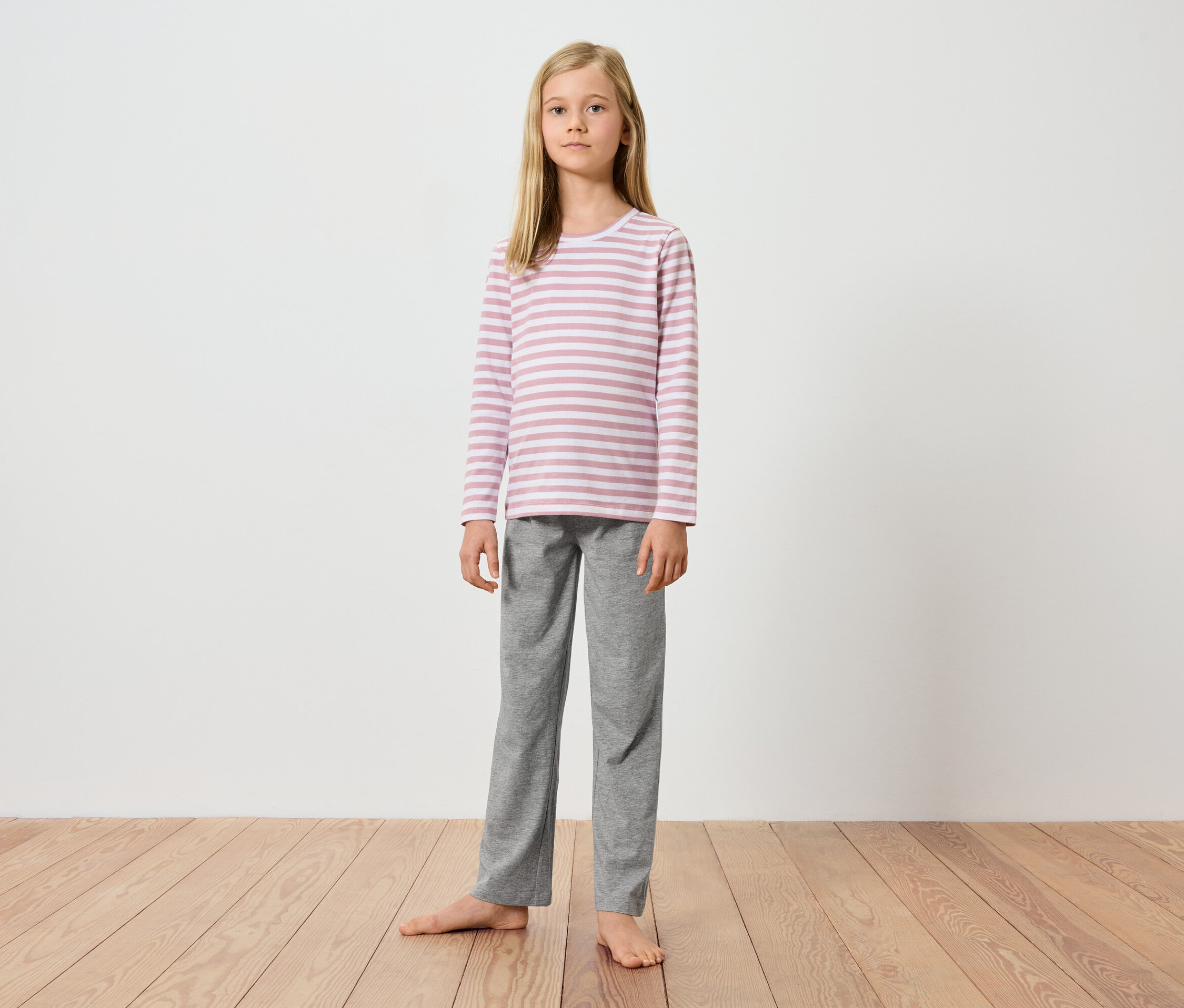 Ein junges Mädchen steht barfuß auf einem Holzboden vor einer weißen Wand. Sie trägt ein langärmeliges, rosa-weiß gestreiftes Oberteil und eine graue Hose.