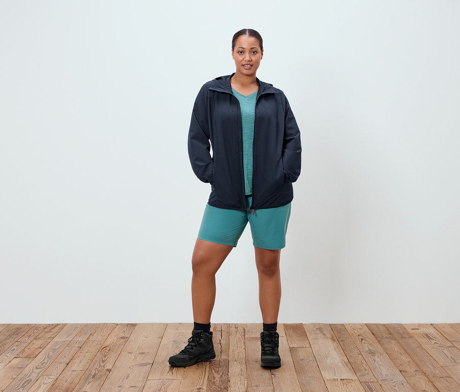 Frau steht mit Händen in den Taschen vor einer weißen Wand. Sie trägt eine marineblaue Jacke, ein türkises T-Shirt, türkise Shorts und schwarze Stiefel.