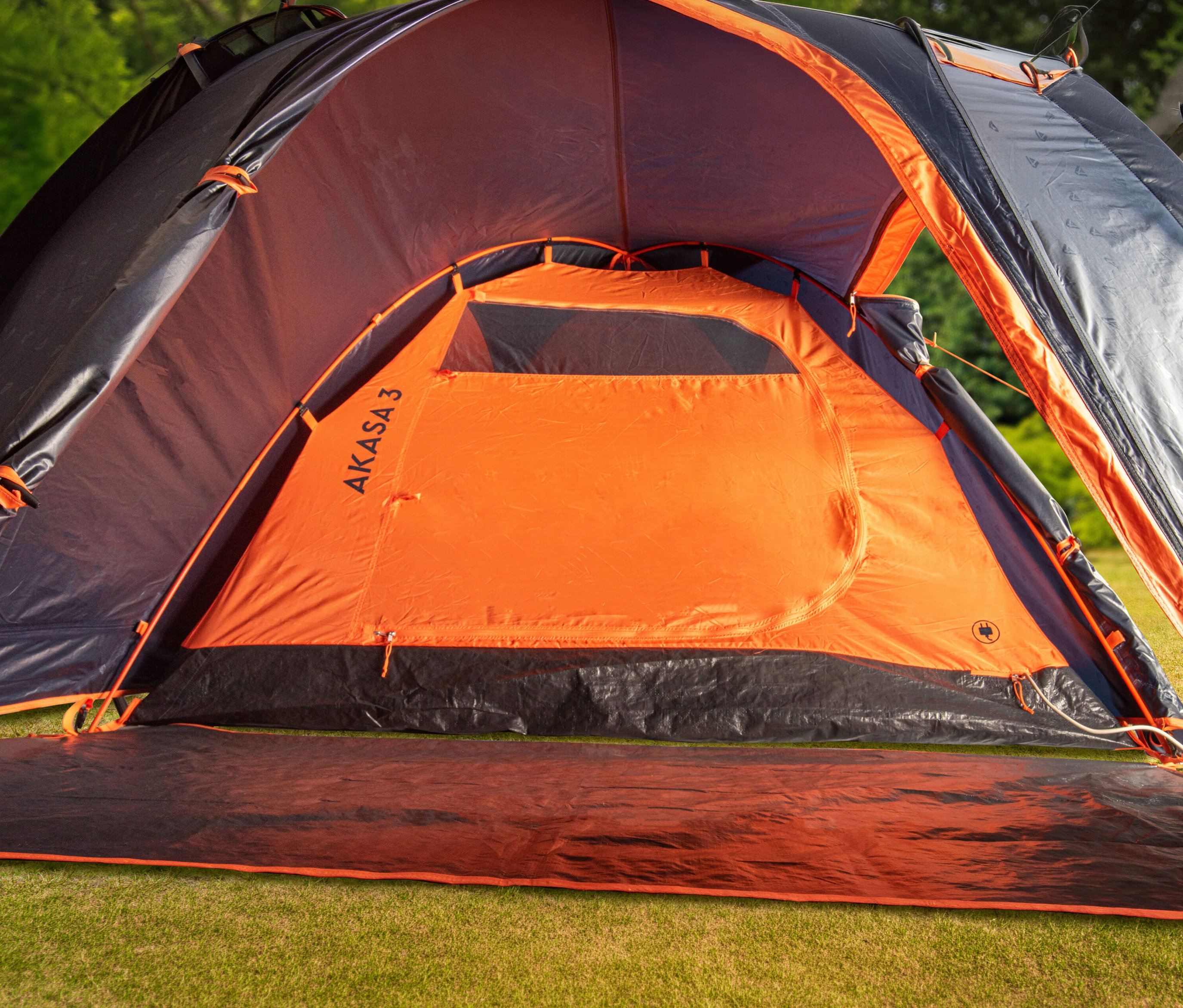 Auf Rasen steht ein offenes, orangefarbenes und schwarzes Zelt mit der Aufschrift AKASA3.