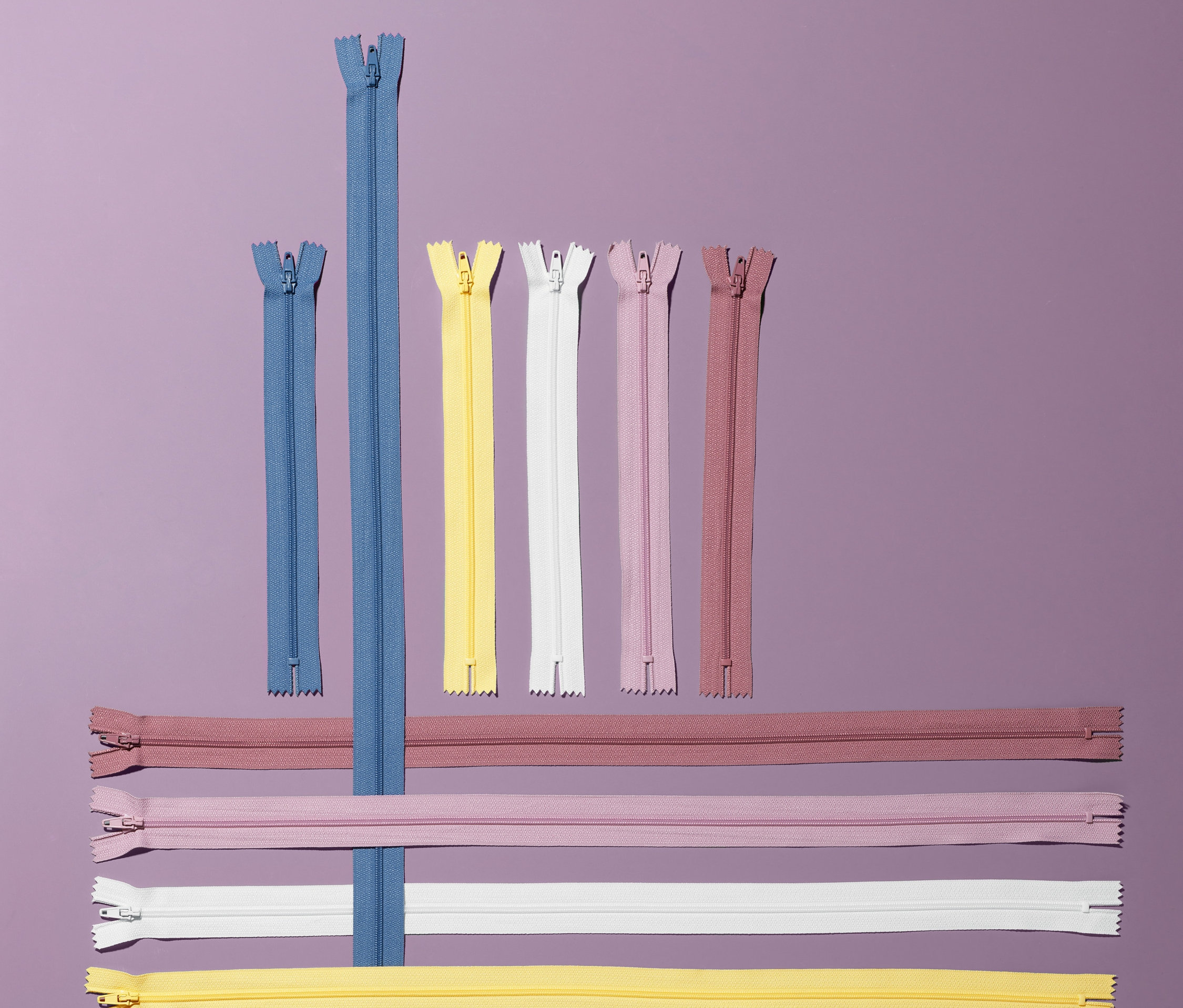 Auf einem lila Hintergrund liegen 10 Reißverschlüsse in verschiedenen Farben und Längen.