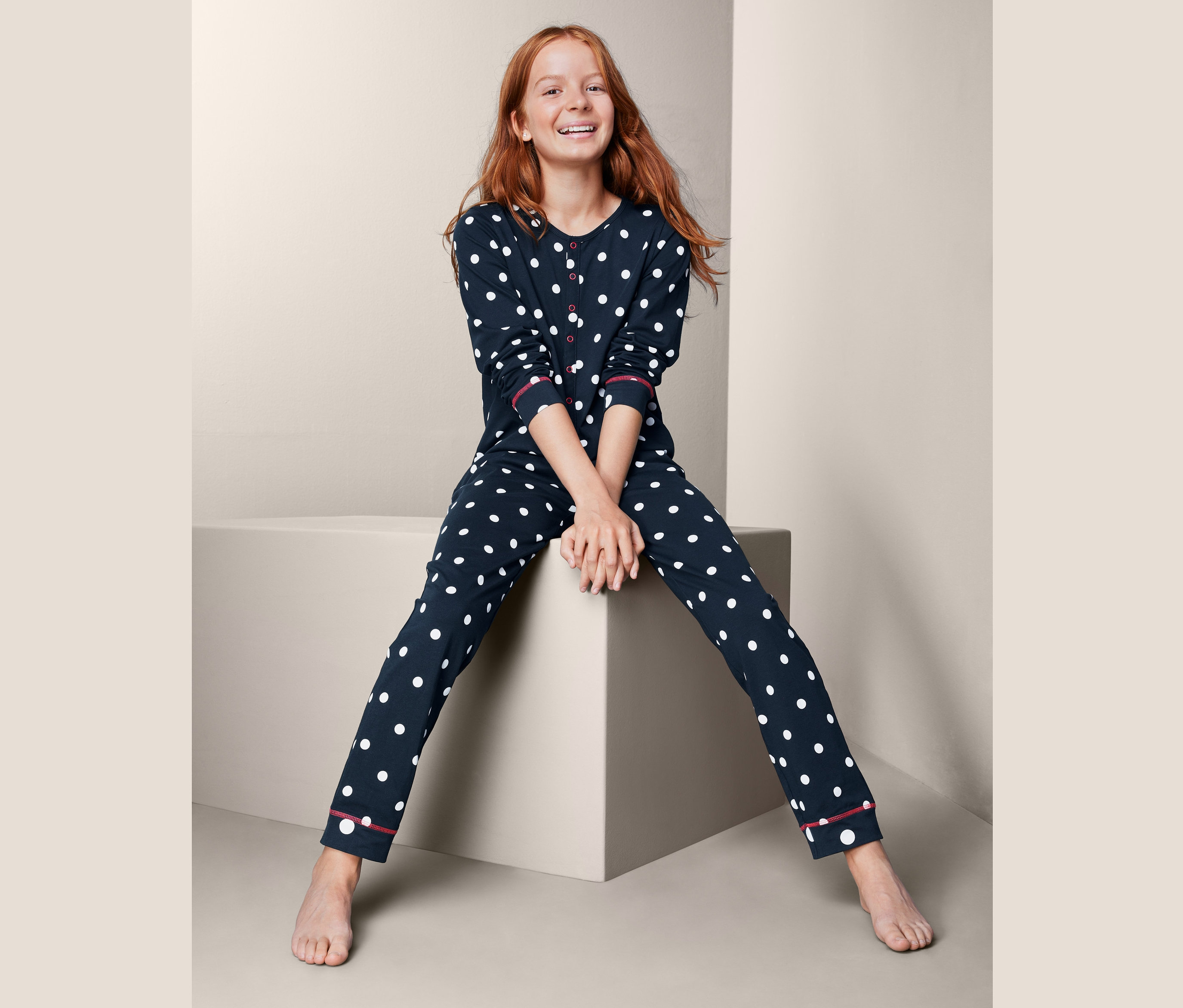 Mädchen sitzt auf einem Würfel und trägt einen marineblauen Kinder-Overall mit weißen Punkten.