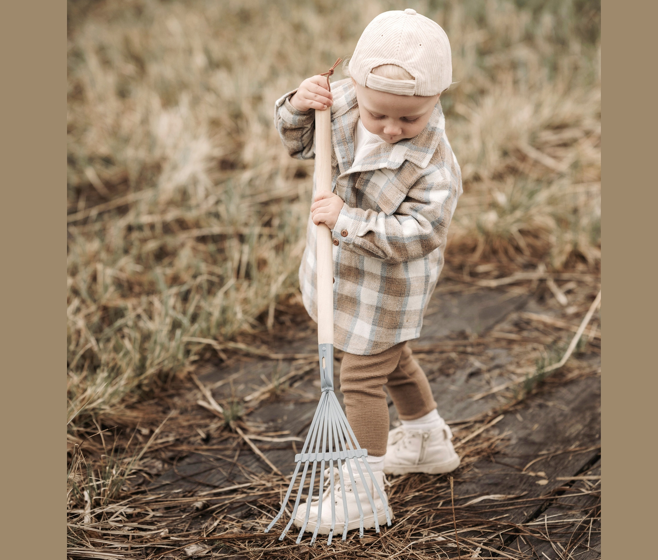 Ein Kind mit kariertem Hemd und Kappe hält ein großes small foot Gartenwerkzeug-Set.