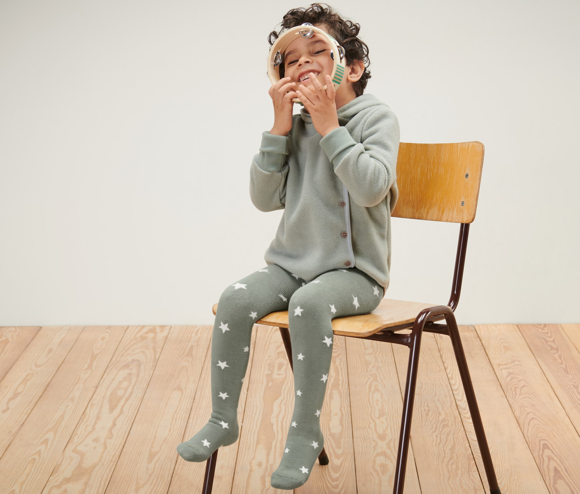 Ein Junge sitzt auf einem Stuhl und spielt mit einem Tamburin. Er trägt eine blaue und grüne Kinder-Thermostrumpfhose.